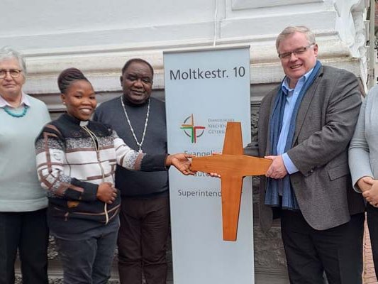Das Altarkreuz aus der Christuskirche in Stromberg – die Kirche wurde im Sommer 2024 geschlossen – findet eine neue Heimat in der Diözese Karagwe, Tansania. Superintendent Frank Schneider (2.v.r.) überreicht es den Gästen Deogratias Aberatha Kokuberwa (3.v.l.) und Benson Boganza bei ihrem Besuch in Gütersloh. Pfarrer Ulrich Klein (links), Christa Marienhagen (2.v.l.) und Maxie Kordes (rechts). 	Foto: KKGT