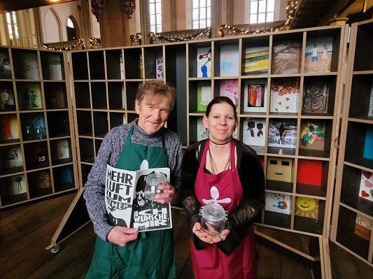 Reinhard Michel und Vanessa Henkelmann begleiten die Kunstaktion. In ihren Händen halten sie Weckgläser, die ab Samstag in der Martin-Luther-Kirche abzuholen sind. Michel und Henkelmann stehen vor dem Regal, das als Suppenregal entstand und im vergangenen Jahr zu einer „Wunschkammer“ wurde.  Aus den Wünschen, die die Gütersloher während der Vesperkirche und im Mai während der „langenachtderkunst“ in dieser Kammer hinterließen, ist ein Heft entstanden, gestaltet von Lara Kees. Gedruckt und gebunden, ist es während der Vesperkirche 2026 zu bekommen.		Foto: privat