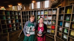 Reinhard Michel und Vanessa Henkelmann begleiten die Kunstaktion. In ihren Händen halten sie Weckgläser, die ab Samstag in der Martin-Luther-Kirche abzuholen sind. Michel und Henkelmann stehen vor dem Regal, das als Suppenregal entstand und im vergangenen Jahr zu einer „Wunschkammer“ wurde.  Aus den Wünschen, die die Gütersloher während der Vesperkirche und im Mai während der „langenachtderkunst“ in dieser Kammer hinterließen, ist ein Heft entstanden, gestaltet von Lara Kees. Gedruckt und gebunden, ist es während der Vesperkirche 2026 zu bekommen.		Foto: privat